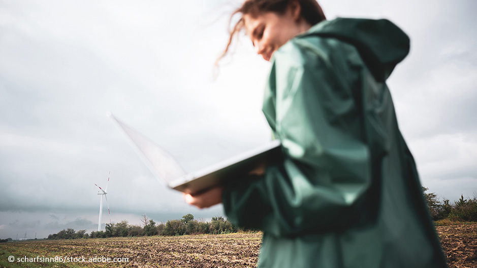 Frau mit Laptop in der Hand steht auf einem Feld mit einem Windrad im Hintergrund